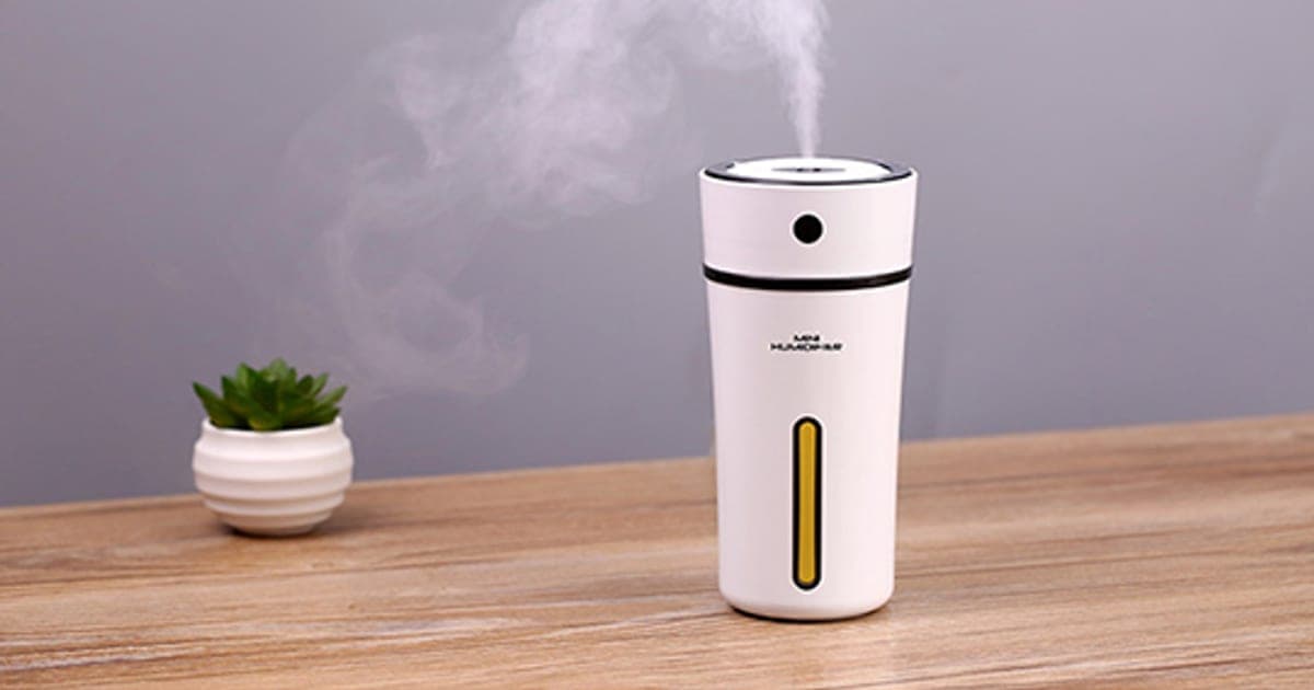White cylindrical AirMoisturize Max mini humidifier on a wooden desk, releasing a plume of mist beside a small succulent