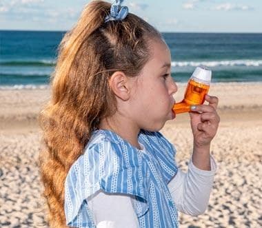 A girl on a beach breathing out through the orange AirPhysio for Children device