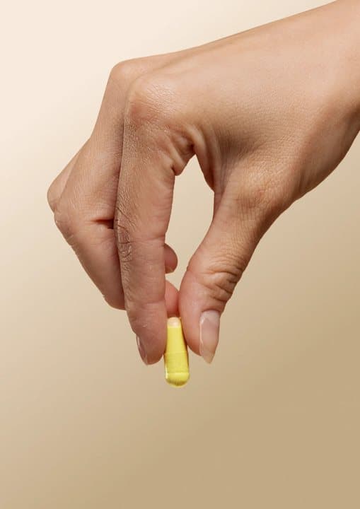 Fingers holding a single yellow Akka capsule against a beige background