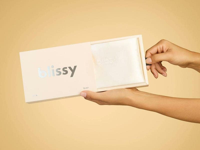 Hands sliding a folded white Blissy silk pillowcase out of its pale pink Blissy gift box