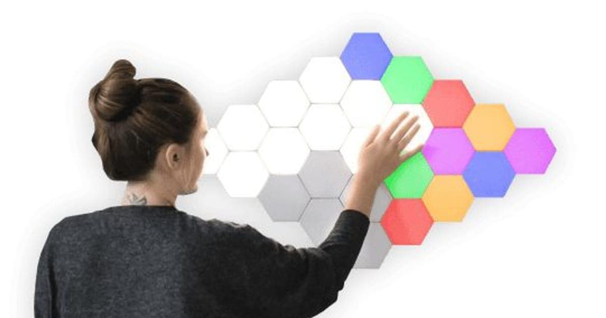 A woman touching a wall of glowing BrightTouch Max hexagon lights in white, blue, green, red, orange and purple