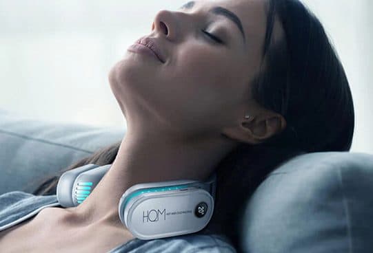 Woman with eyes closed resting her head back on a sofa, wearing a white horseshoe neck cooler with blue lights and a temperature display