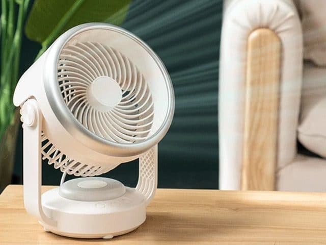 White CoolSwing fan with a silver rim and curved white stand on a wooden table, plant and armchair behind