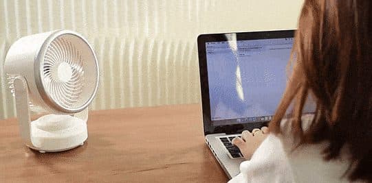White CoolSwing air-circulator fan on a wooden desk facing a person with long hair working on a laptop