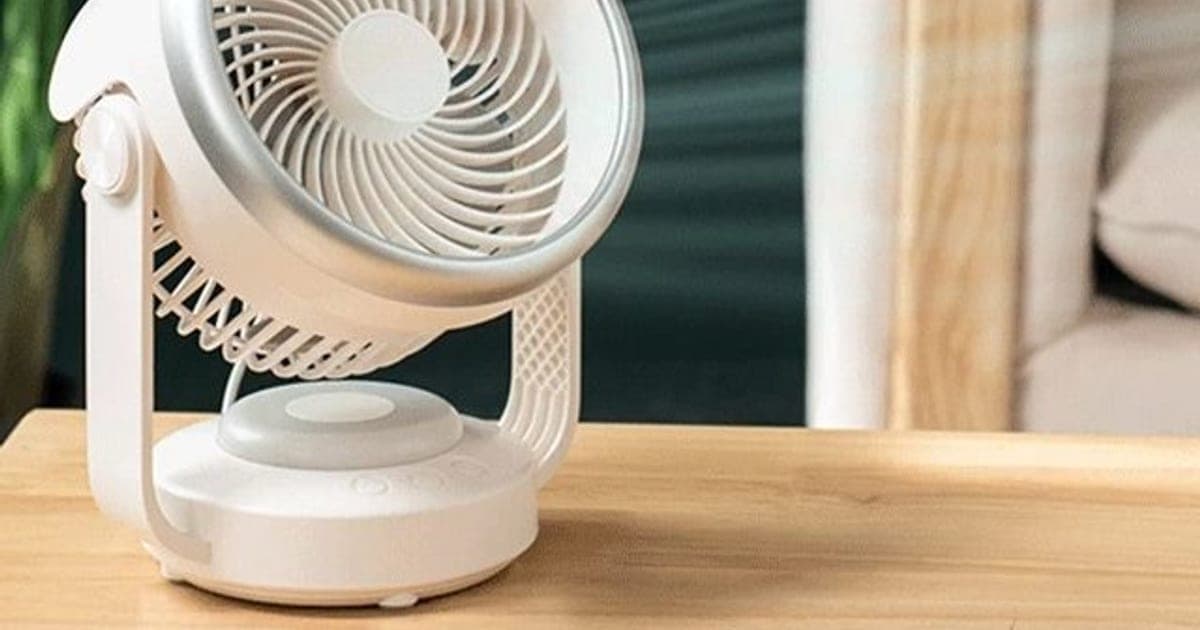 White round CoolSwing desk fan with a silver rim, spiral grille and curved stand on a wooden table, with a plant and armchair behind