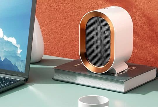 White CozyTime Pro compact heater with a copper-coloured oval rim and mesh grille, on a hardback book next to a laptop and a mug