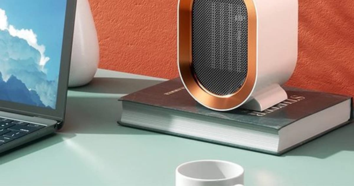 White and copper-trimmed CozyTime Pro oval heater standing on a book on a green desk beside a laptop, against an orange wall