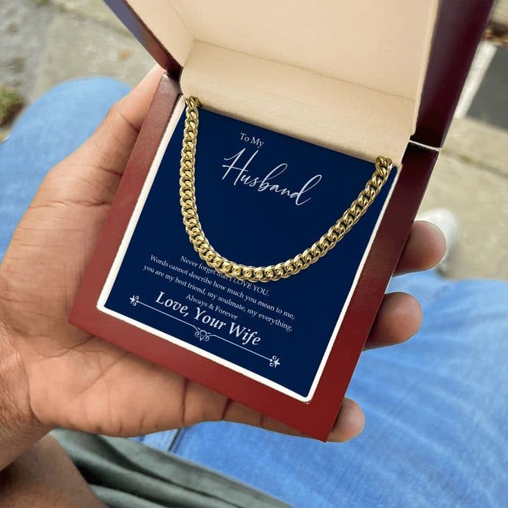 Hand holding the gold-tone Cuban Link Chain on its To My Husband card in an open mahogany-style box, above a pair of jeans