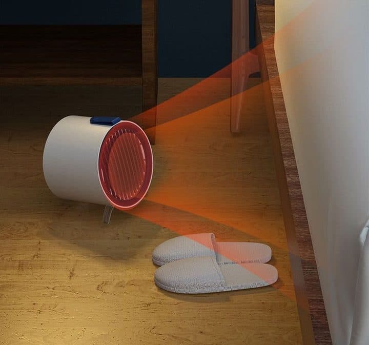 EcoThermal heater on a wooden bedroom floor beside a bed, glowing red and beaming warmth towards a pair of slippers