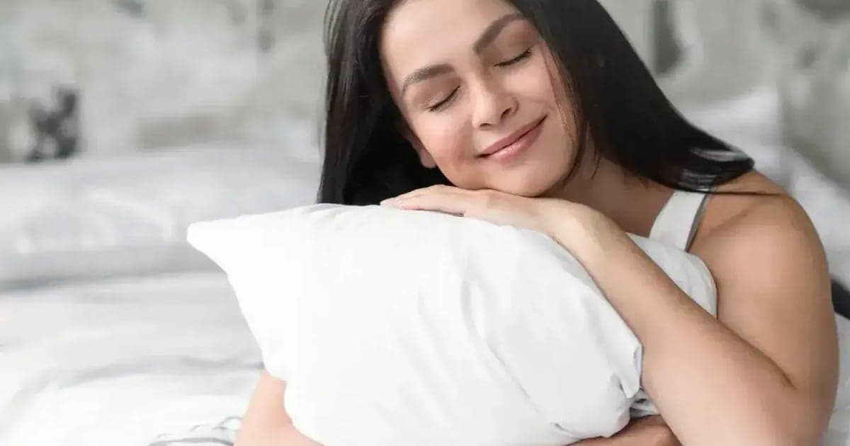 A woman with her eyes closed hugging a plump white FluffCo pillow in a bed with white bedding