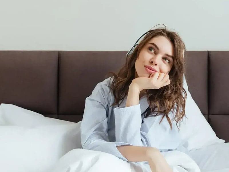 A woman sitting up in bed propped against white FluffCo pillows in front of a dark headboard
