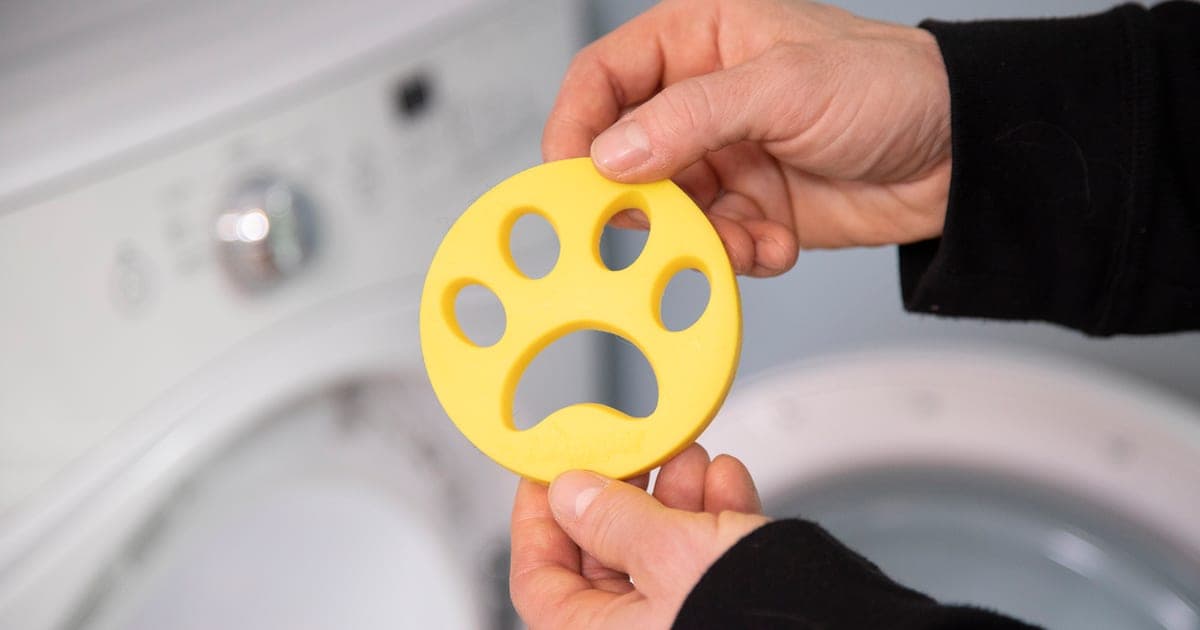 Hands holding a yellow paw-shaped FurZapper disc in front of a front-loading washing machine