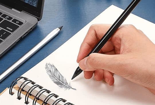 Hand holding a black FutureX Pen over a spiral notebook with a feather drawing, next to a silver pen and a laptop