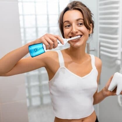 Woman in a bathroom smiling as she uses the JetFloss Pro with a blue tank, holding a model tooth