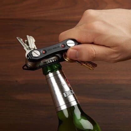 Hand using a black key organizer marked Key Ninja to open a beer bottle, with keys folded in