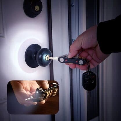 Person using a key organizer with a lit LED to find a door lock in the dark, with an inset of the torch on a table