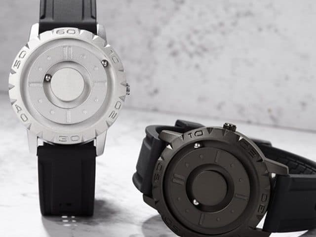 Silver and gunmetal MagWatch models side by side on black straps against a pale stone background