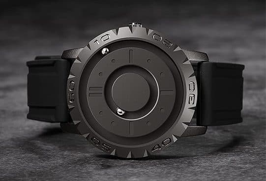 Dark grey MagWatch lying on a stone surface, with raised hour markers, a central disc and two small steel balls on its inner and outer tracks