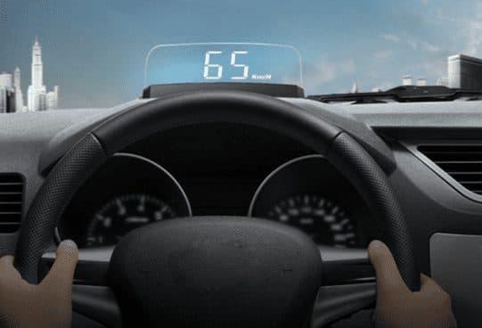 Hands on a steering wheel with a MasterHUD Pro on the dashboard projecting 65 km/h in white digits onto a clear panel