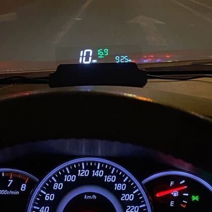 Night view through a windscreen with a head-up display showing speed, voltage and time in coloured digits above the instrument cluster
