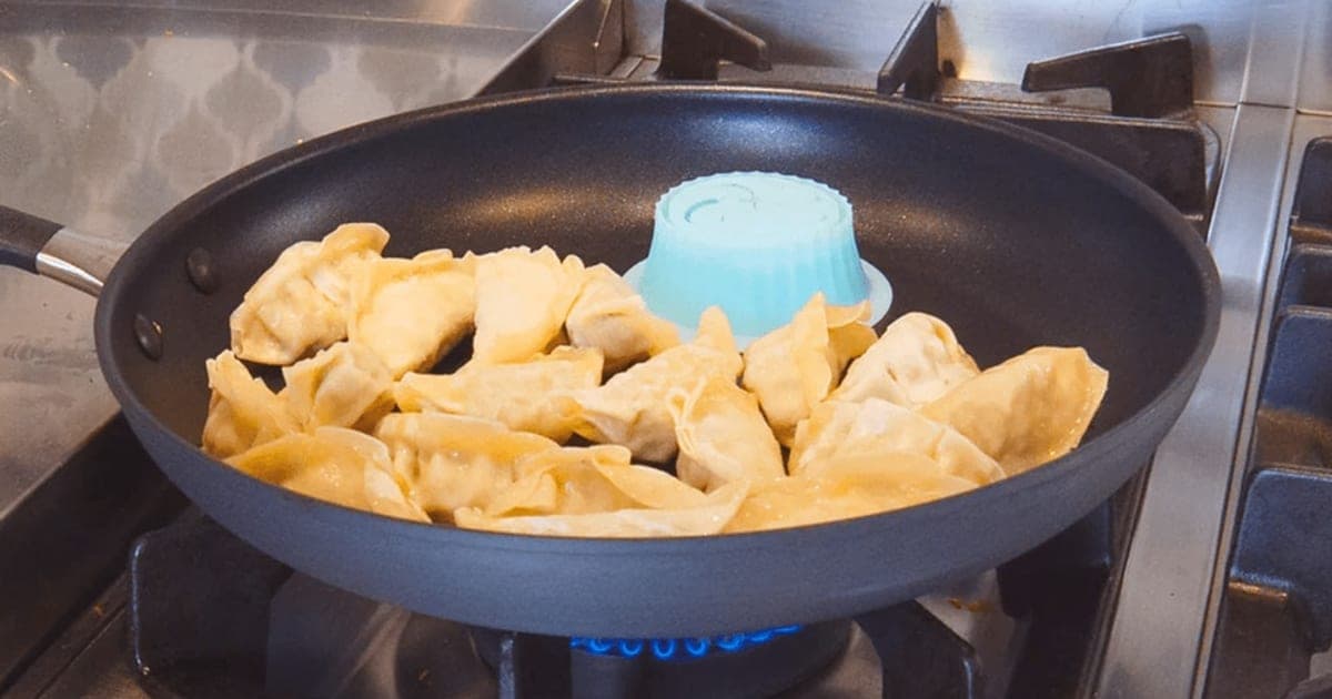 Blue Pacuzzi silicone steamer cup in a frying pan surrounded by browned dumplings on a gas hob