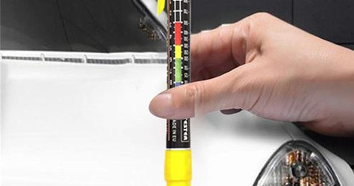 Hand holding the PaintPen Pro gauge with a yellow tip against a white car bonnet, its scale showing green, yellow and red bands