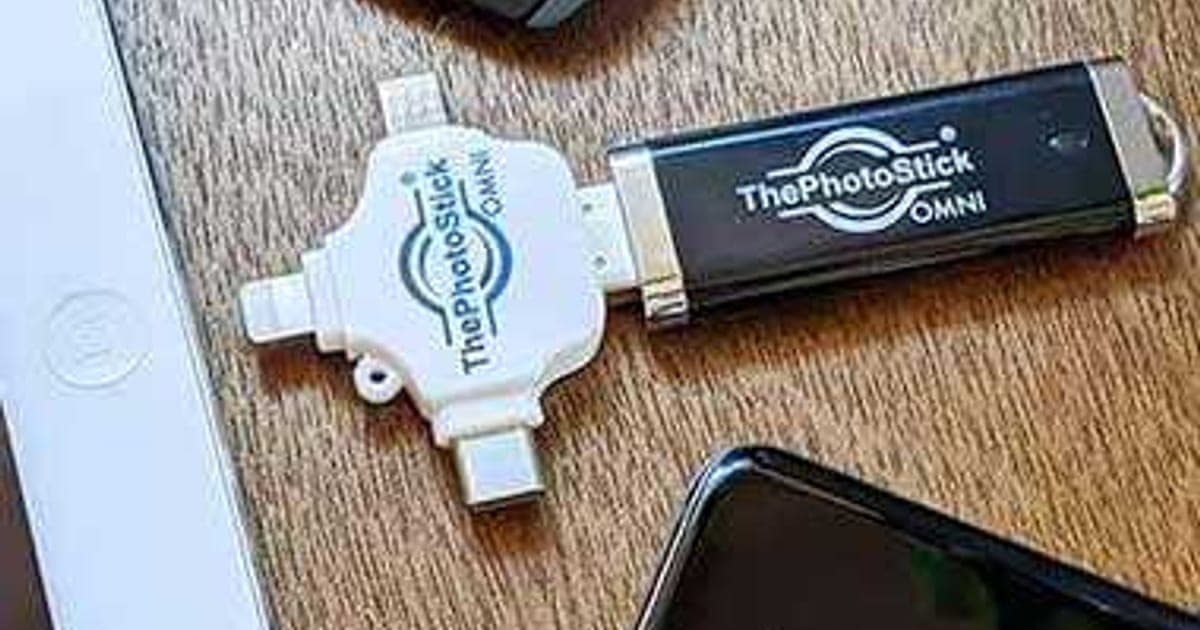 ThePhotoStick Omni with its white four-way adapter attached, lying on a wooden table beside a tablet and phone