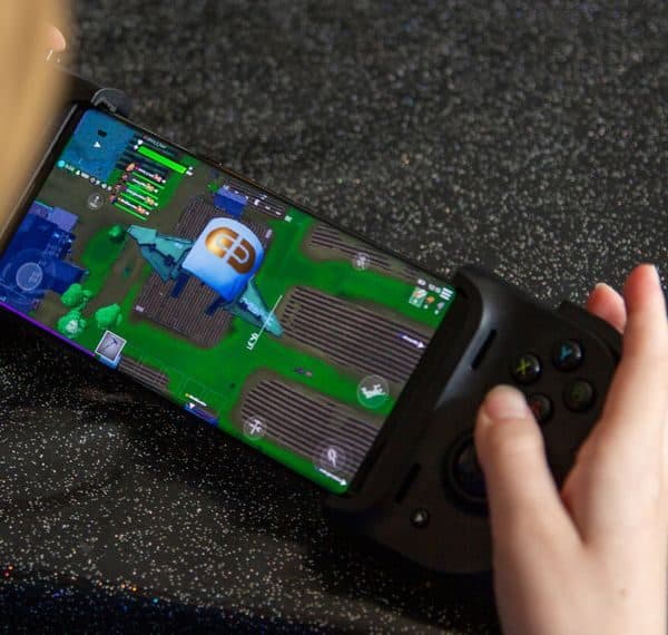 Hand on the right grip of a compact black controller attached to a phone running a colourful battle royale game on a sparkly surface