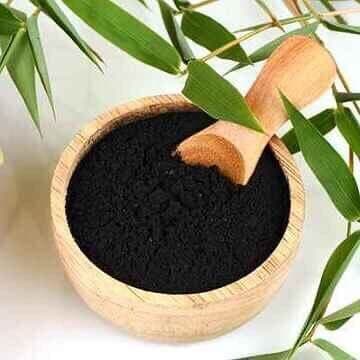 Wooden bowl of fine black charcoal powder with a wooden scoop and green bamboo leaves, used for the premium bamboo charcoal add-on