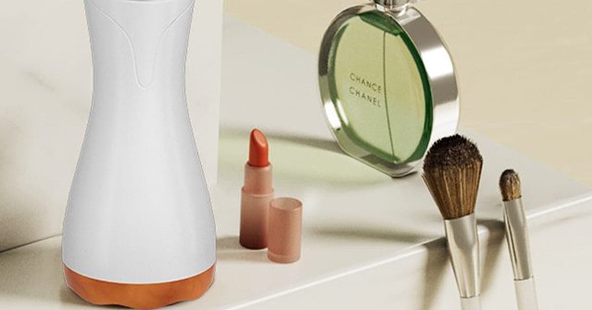White hourglass-shaped RelaxMe Pro massager with a copper-coloured base and a digital top display, on a dressing table beside make-up brushes and a perfume bottle