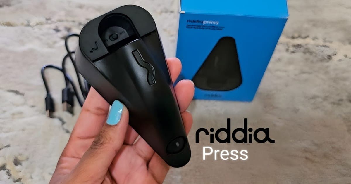 Black riddiaPress cordless mini iron held in a hand next to its blue box