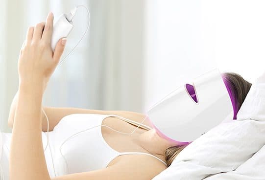 Woman relaxing in bed with the white SkinRevive Pro mask over her face, purple light at the eye slot, holding a white handheld controller on a cable
