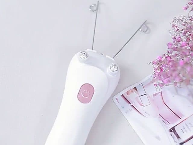 White SoftEpilator Pro with wire arms and a pink button lying beside dried pink flowers