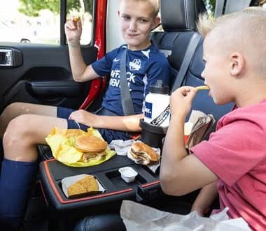 Two boys in the back seat eating burgers and fries on a Stupid Car Tray placed between them