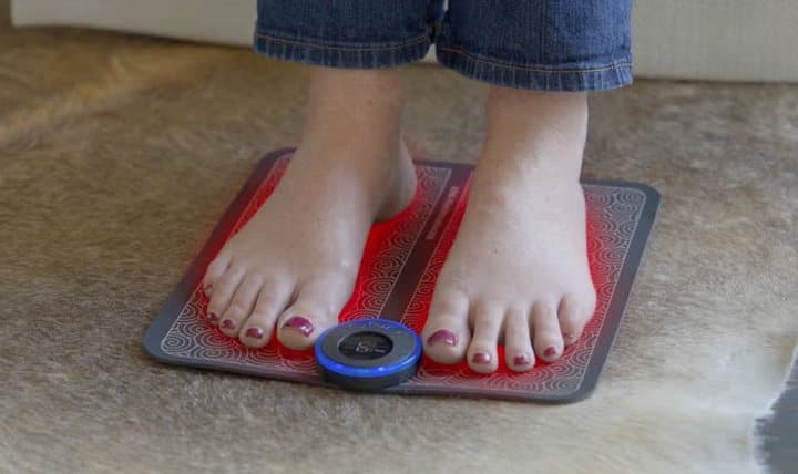 Bare feet resting on a TheraFoot Pro pad with a glowing red outline and blue controller on a rug
