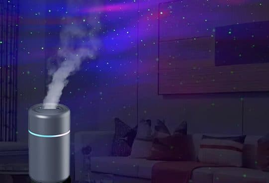 Small grey cylindrical TwilightMist humidifier misting on a floor in front of a sofa, with a starry purple galaxy pattern across the walls