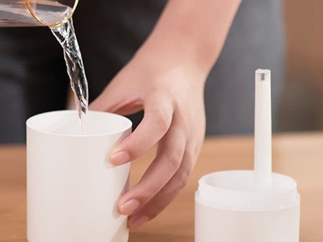 Hand pouring water from a glass jug into the white TwilightMist base, with the lid and wick standing beside it