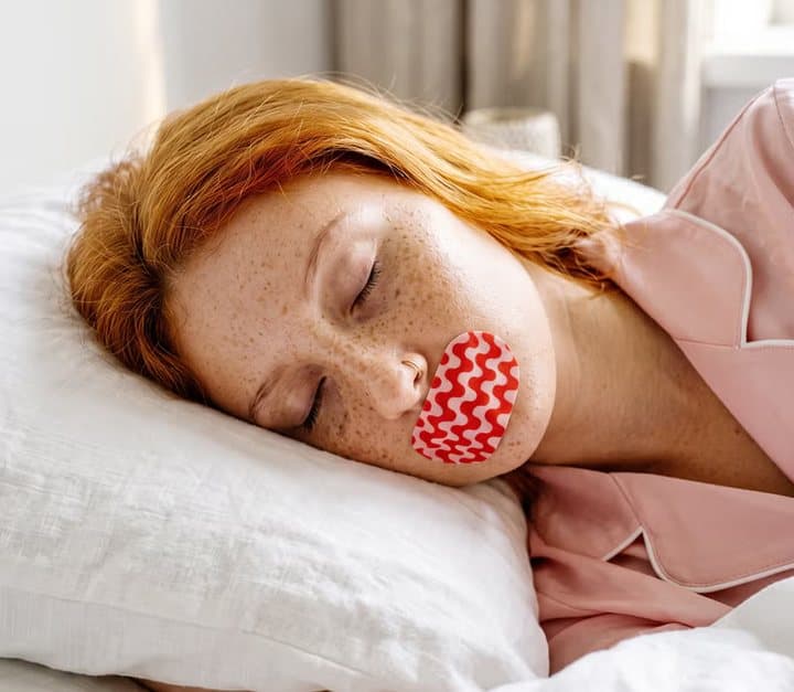 Woman asleep on a pillow wearing a red patterned Ugly Sleep Club strip over her lips