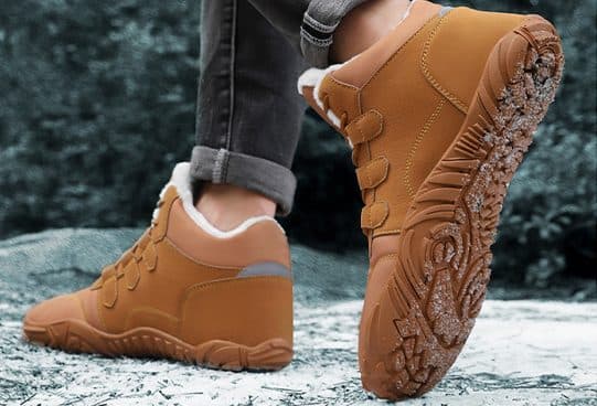 Person wearing tan fleece-lined WinterGlide Pro boots with three straps and jeans, on frosty ground, one sole with snow in the lugs