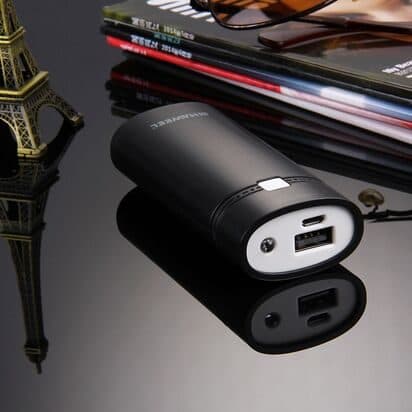 Black cylindrical power bank case with a USB port and a light, on a reflective table beside magazines