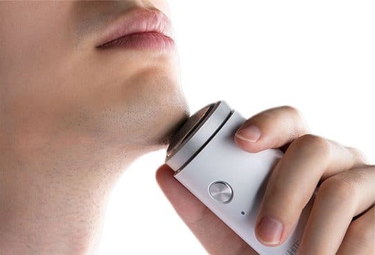 Close-up of a man's chin and lips with a white mini ZeroShave Pro shaver pressed to the stubble, showing its single round head and button