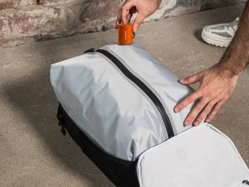 A hand holding the orange Aerless pump on the valve of a packed grey vacuum bag inside an open backpack
