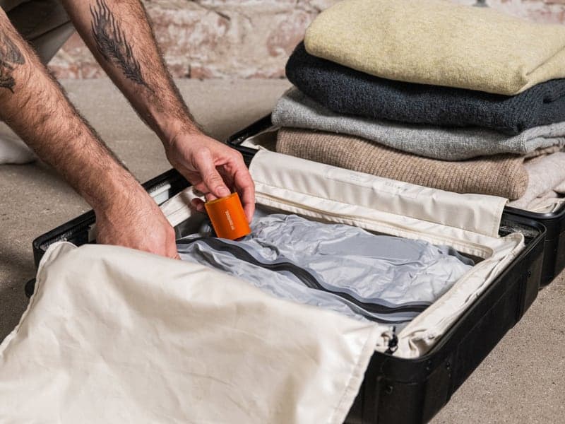 Compressed grey Aerless bags lying flat in an open suitcase, with the orange pump on top and folded sweaters beside it