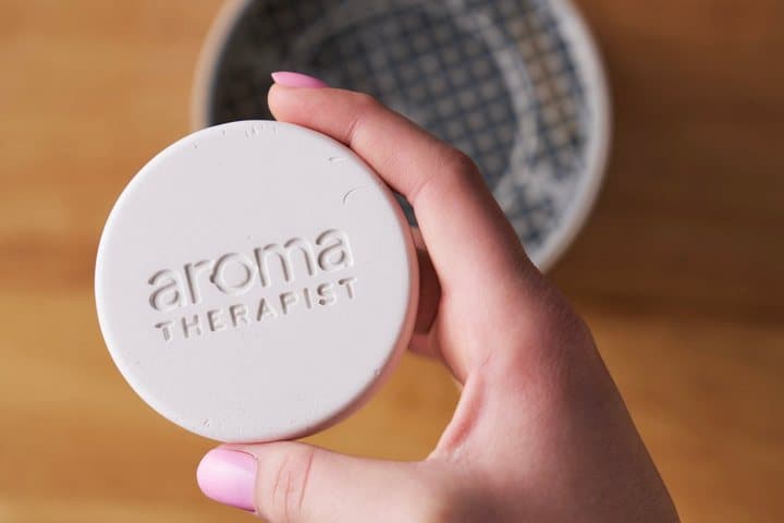Hand holding a white AromaPuck disc embossed with the Aroma Therapist logo above the Amplifier