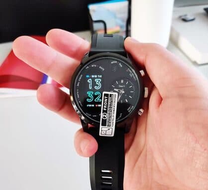 Hand holding a BiT Watch with a protective sticker on the face, showing 13:32 on the digital screen beside the analogue sub-dial