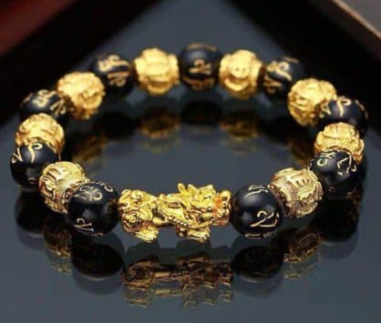 Bracelet of black carved beads alternating with gold patterned beads and a gold Pixiu, on a dark reflective surface