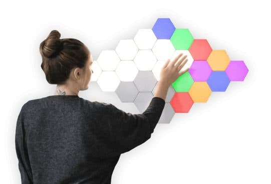 A woman with her hair in a bun touching a large cluster of hexagon wall lights, some white and some lit blue, green, red, orange and purple