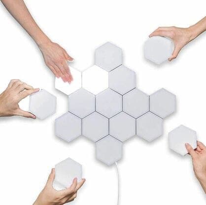 Several hands arranging white hexagon light panels into a cluster on a white surface, one panel lit