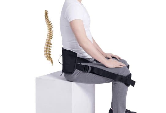 Side view of a seated man in grey trousers wearing the CorrectBack black lumbar pad with two straps running to his knees, with a spine model beside him