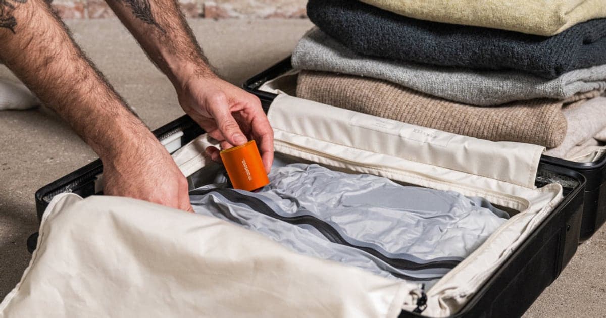 A hand holding a small orange pump over a compressed grey vacuum bag in an open suitcase, beside a stack of folded sweaters